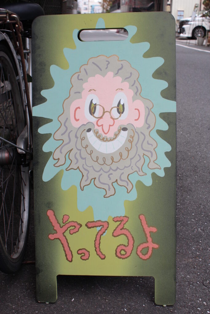「やってるよ」のメッセージと、ひげをはやした笑顔の男性のイラストがかかれた立て看板